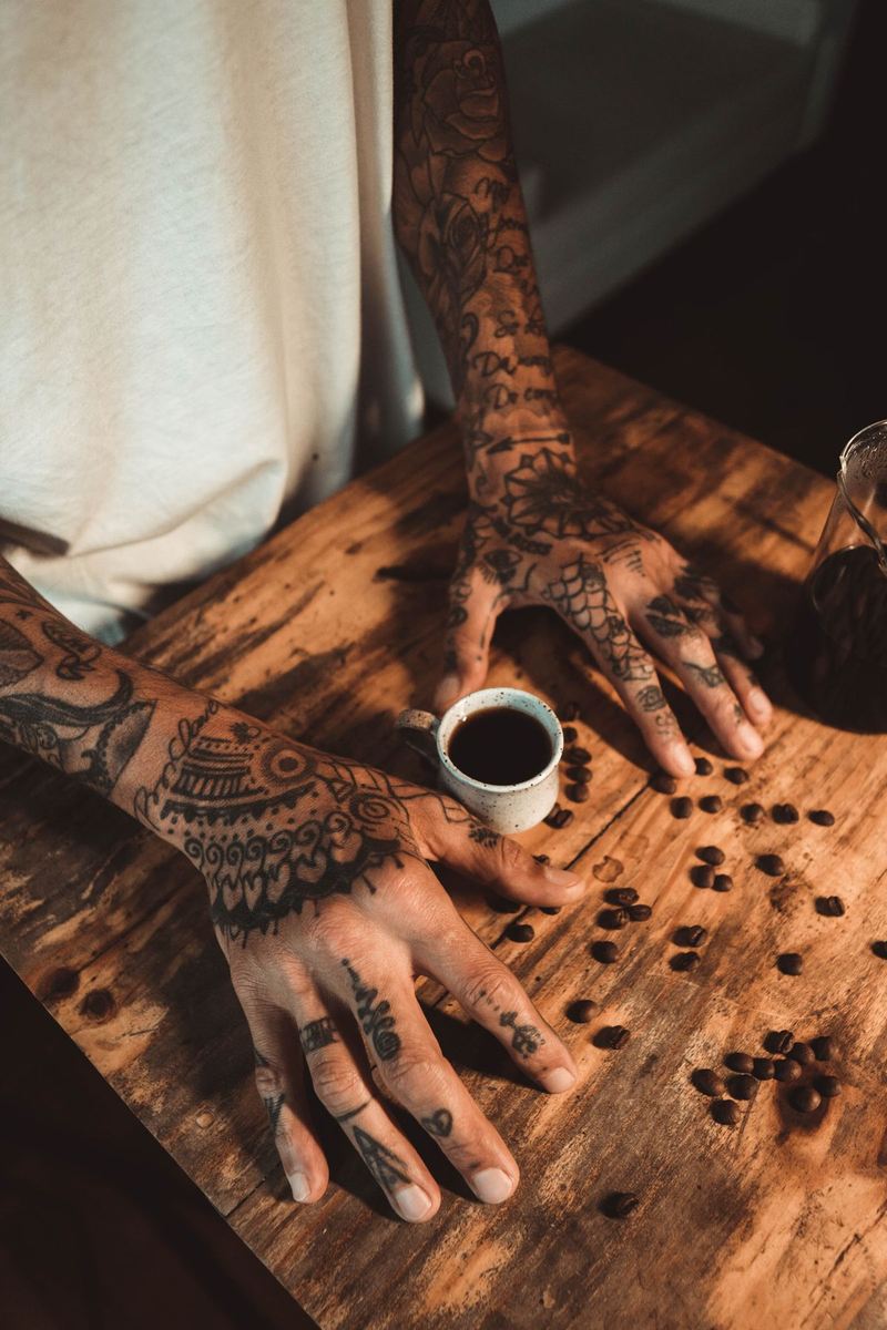 Tattooed hands holding an espresso at a wooden café table