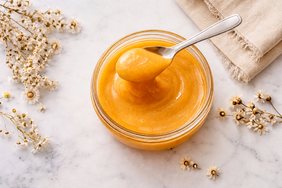 Creamed honey in a glass bowl with a silver spoon, small white flowers and neutral linen.